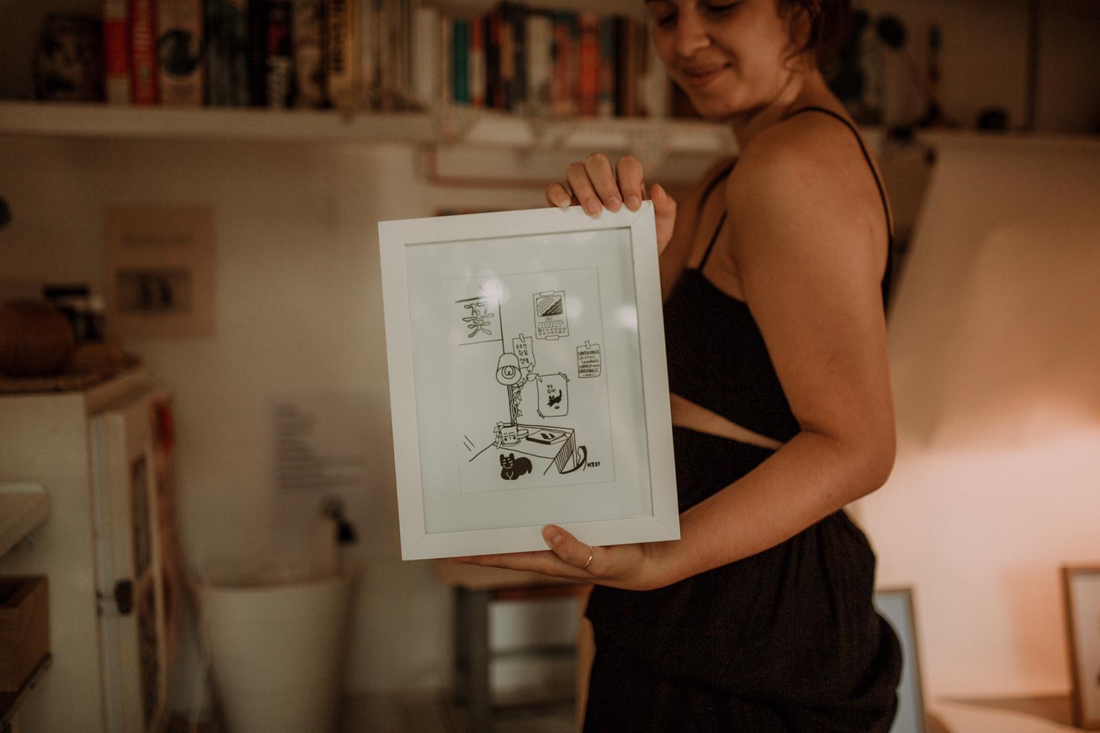 Kikitsa Draws, inside caravan, holding framed “Cat Resting in Flower-café”